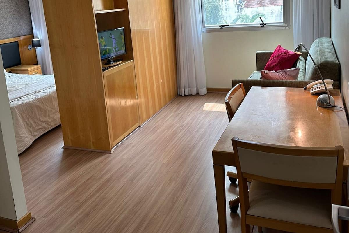 A hotel room with a wooden wardrobe, a bed, a TV, a dining table with chairs, a sofa, and a window with curtains.