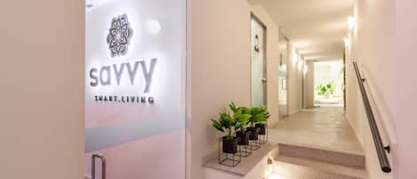 Una entrada moderna de hotel con puerta de vidrio que muestra el logotipo y el nombre 'savvy smart living'. Se ve una escalera con barandilla y plantas en macetas.