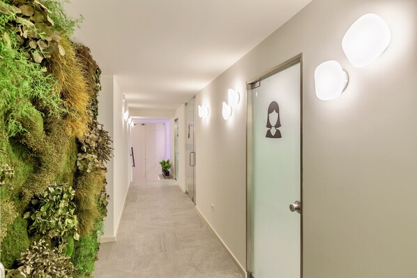 A hallway with a green wall installation, a door with a person silhouette, and wall-mounted lights.