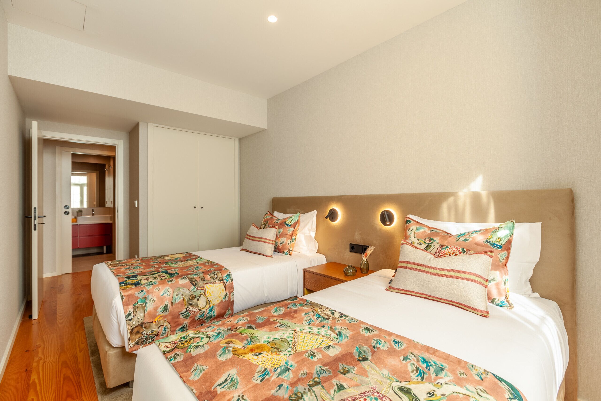 A hotel room with two beds, a wooden floor, a bathroom with a red sink, and a hallway leading to another room.