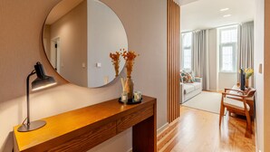 Una habitación de hotel moderna con una consola de madera, un espejo redondo, una lámpara de escritorio, un jarrón con flores secas y vistas al área de estar contigua.