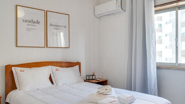 A neatly made bed with white linens and towels, a wooden headboard, a nightstand with a lamp, and two framed signs on the wall. - Almada Saudade by Hopstays | Mezzanine & Charm (Almada)
