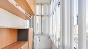 A narrow balcony with a wooden shelf, a white radiator, and a view of buildings through vertical blinds. - Almada Saudade by Hopstays | Mezzanine & Charm (Almada)
