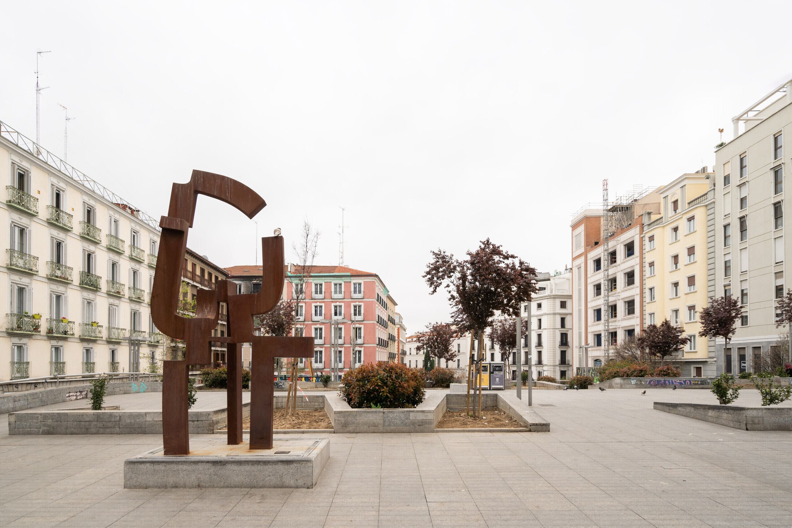 A public square with a large metal sculpture, surrounded by multi-story buildings with balconies and windows.