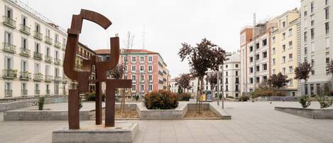A public square with a large metal sculpture, surrounded by multi-story buildings with balconies and windows.