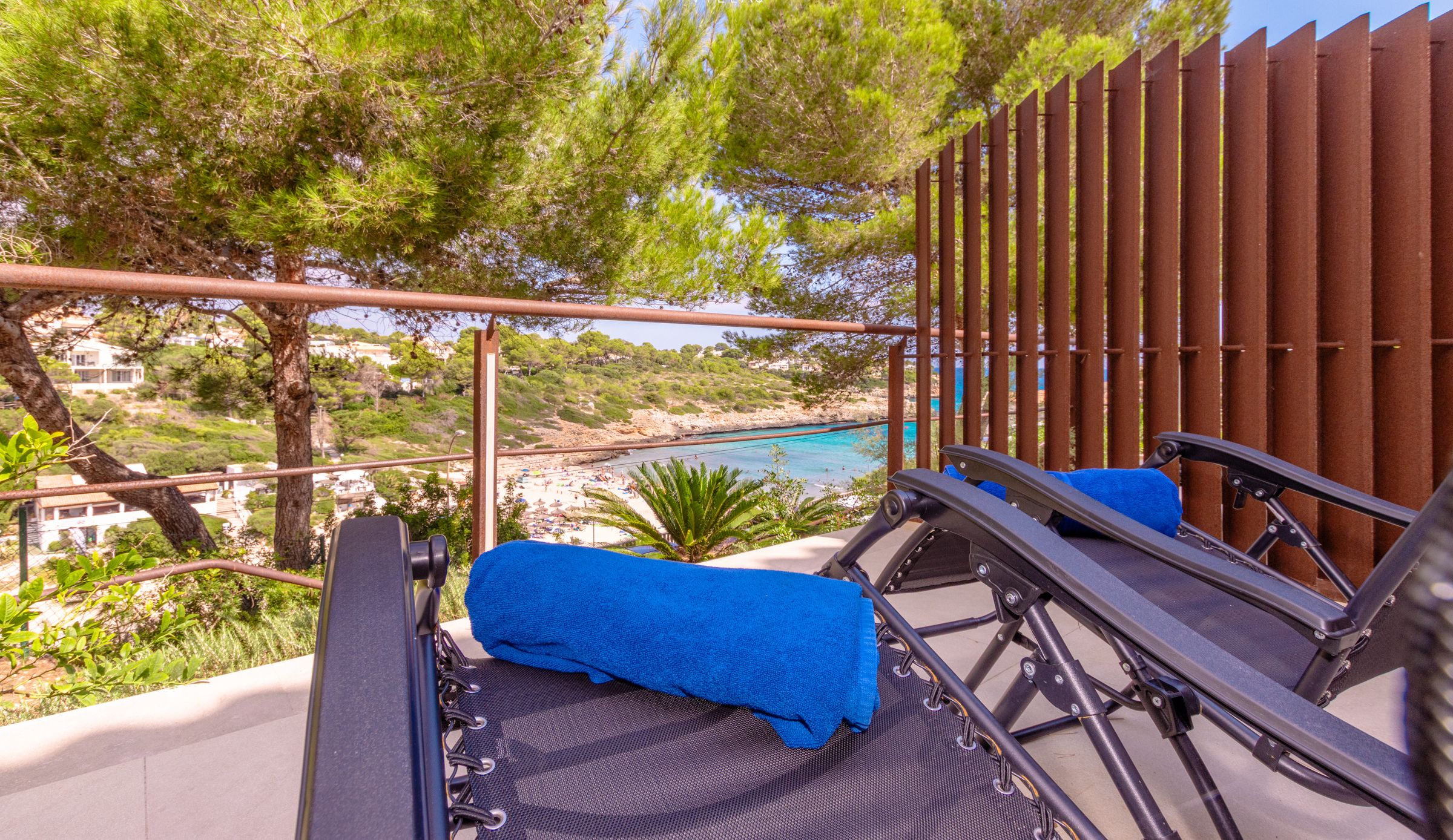 A balcony with a view of a beach and turquoise waters. There are two lounge chairs with blue cushions.