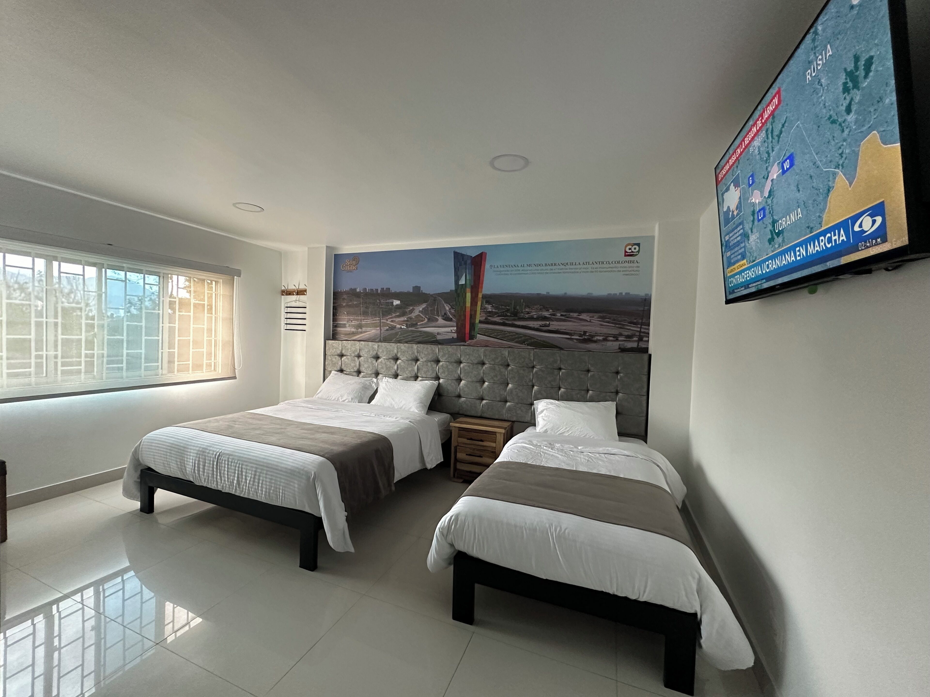 A hotel room with two beds, a large window, and a wall-mounted TV displaying a map.
