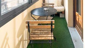 A narrow balcony with a round table, two wooden chairs, a woven light fixture, a sofa with cushions, and a view of buildings through large windows. - Bamboo Caban 2 min du centre ville (Annecy)