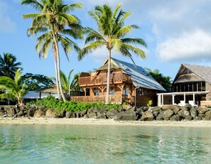A wooden house with a balcony, situated by a beach with clear water and palm trees. - Koromiri Bungalow (Rarotonga)