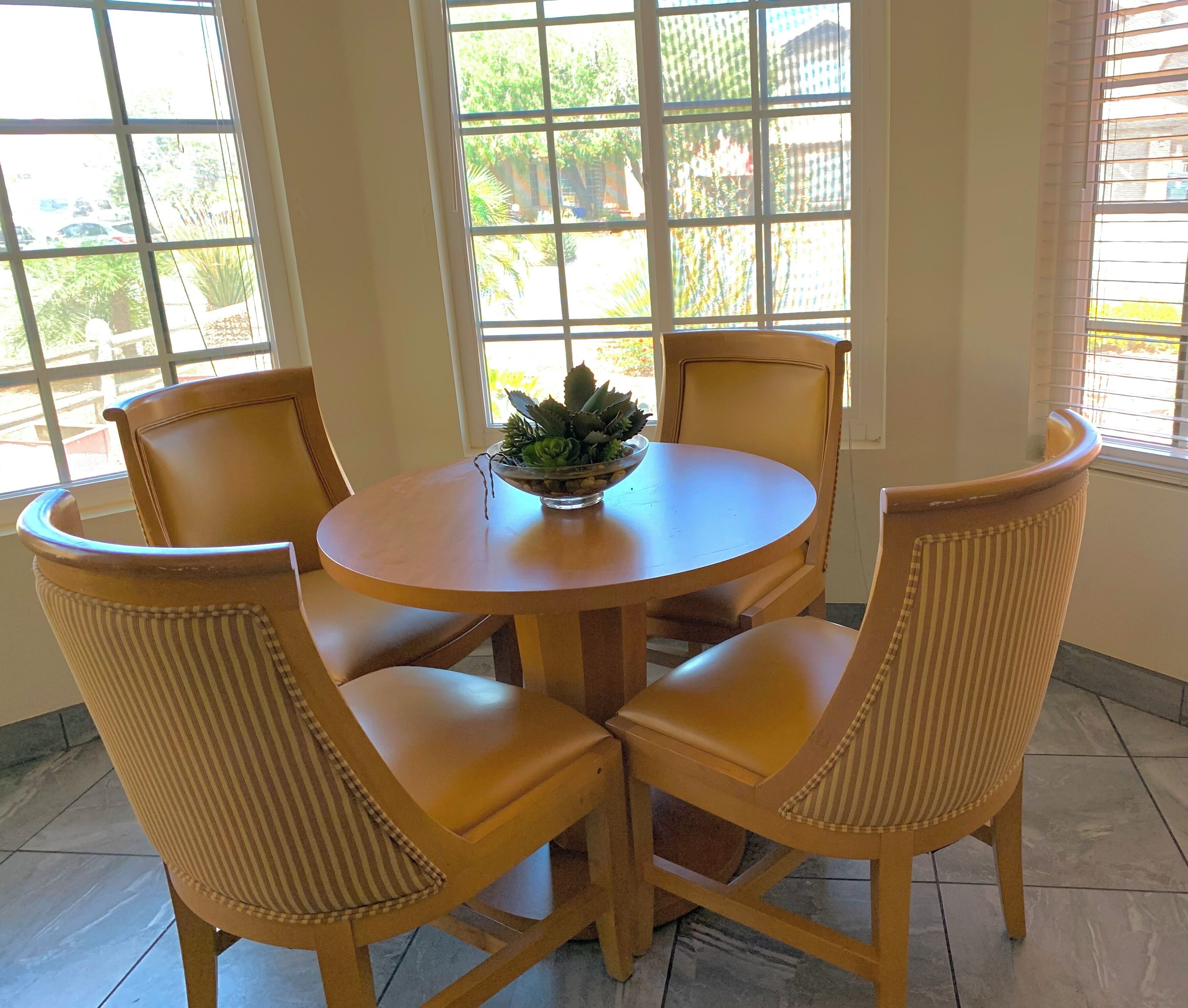 Una mesa de madera redonda con un cuenco de plantas, rodeada por cuatro sillas a rayas en una habitación luminosa con ventanas grandes.
