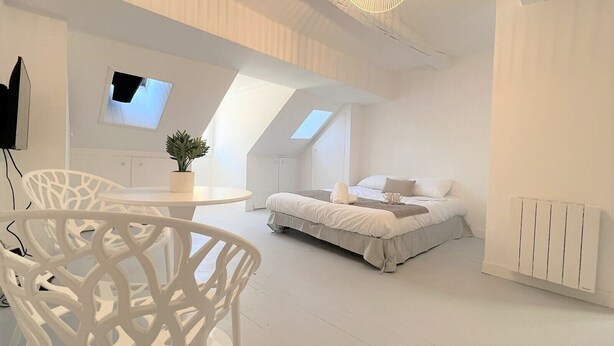 Un dormitorio moderno con decoración blanca, una cama, una mesita y una silla.
