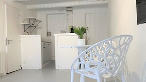 Un interior moderno y luminoso en tonos blancos, con una zona de comedor que incluye una silla blanca y una mesa. Se observa también una cocina con un refrigerador y una planta en maceta.