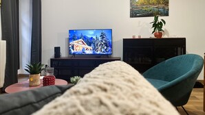 Ein Wohnzimmer mit Fernseher, der eine verschneite Landschaft zeigt, einem türkisfarbenen Sessel, einem kleinen Tisch mit Pflanze und roter Tasse sowie einem Gemälde an der Wand.