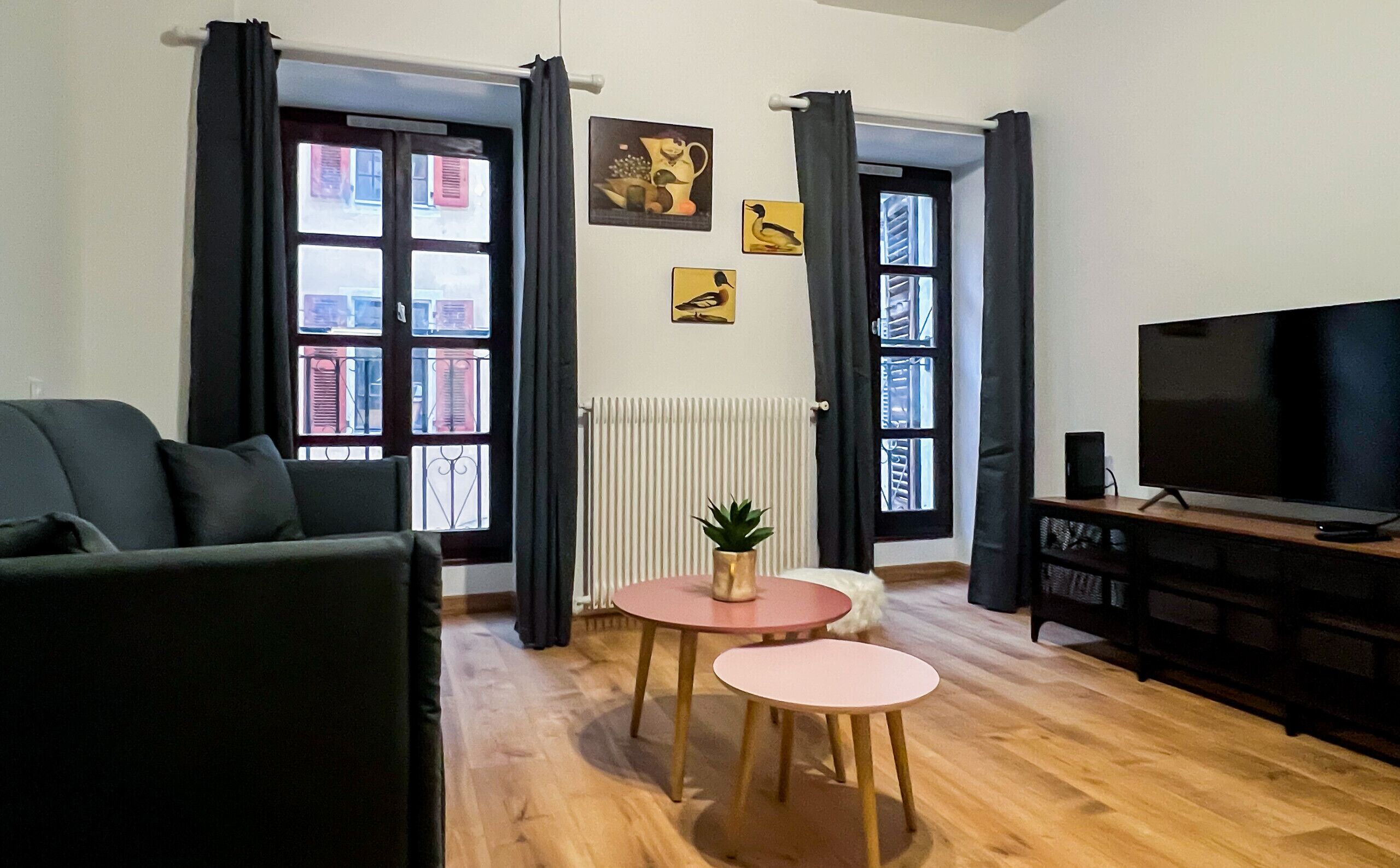 Un salon avec un canapé, deux petites tables, un téléviseur et de grandes fenêtres avec des rideaux.