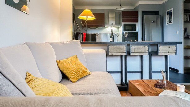Ein Wohnzimmer mit grauem Sofa, gelben Kissen und einem Holztisch. Im Hintergrund ist eine Küche mit einer gelben Lampe und modernen Geräten zu sehen.