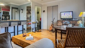 Ein modernes Wohnzimmer mit Couch, Esstisch und Stühlen. Ein Couchtisch aus Holz sowie ein Fernseher auf einem Schrank und eine Gitarre, die an der Wand lehnt, sind ebenfalls zu sehen.