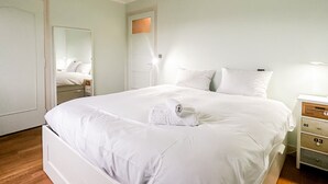 Ein ordentlich bezogenes Bett mit weißen Leinen und einem aufgerollten Handtuch, ein Nachttisch mit Schubladen und ein Spiegel an der Wand.