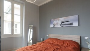 A bedroom with a bed, a bedside table, a nightstand, a mirror, and a window with blinds.