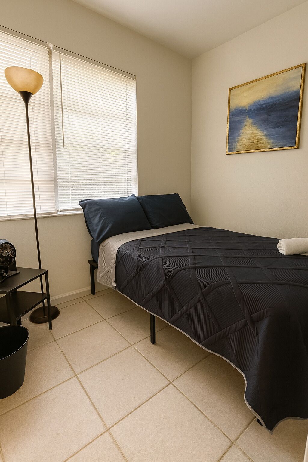 A bedroom with a bed, a standing lamp, a picture on the wall, and a window with blinds.