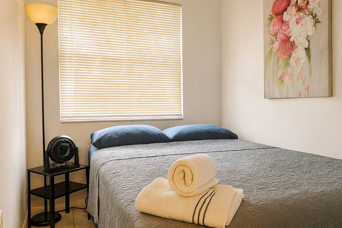 Un dormitorio con cama, mesita de noche, lámpara, ventana con persianas y un cuadro en la pared.