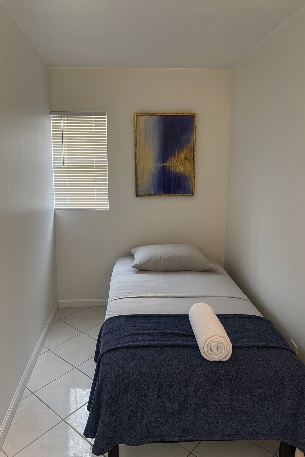 Una habitación pequeña y sencilla con cama, ventana con persianas y un cuadro en la pared.