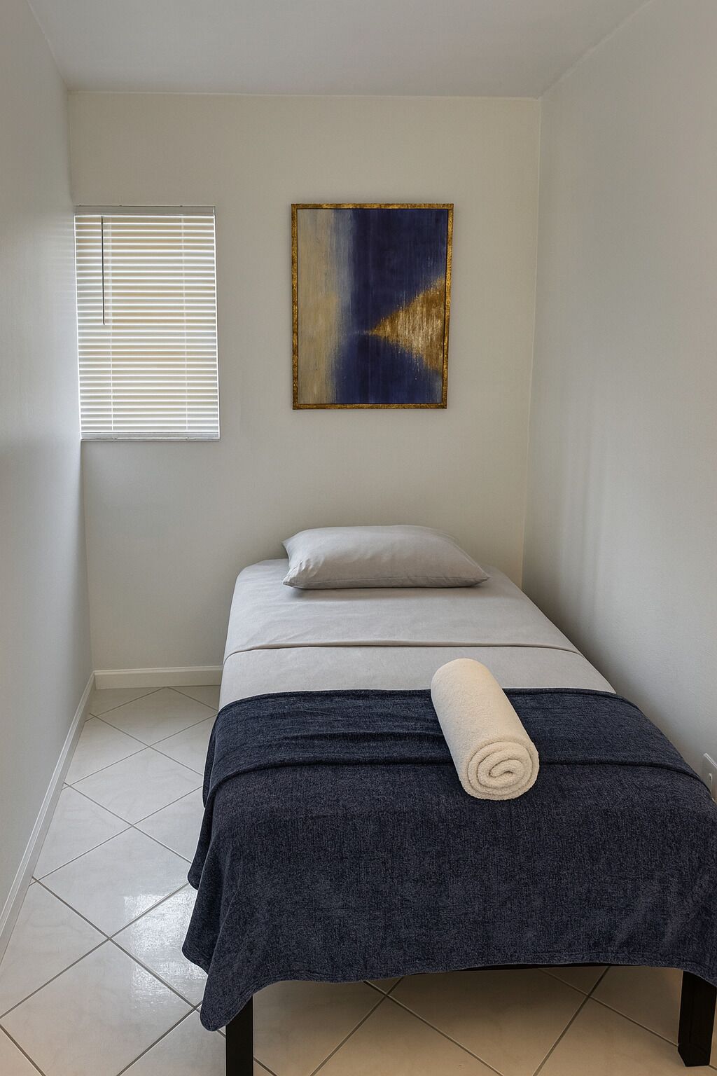 A small, neatly arranged bedroom with a bed, a pillow, a rolled towel, and a painting on the wall.