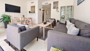 Ein modernes Wohnzimmer mit einem grauen Sofa, einem Esstisch mit Stühlen, einem Flachbildfernseher und einer Vitrine mit Dekorationsobjekten.