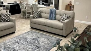 A modern kitchen with dark cabinets, a stainless steel refrigerator, and a dining area with a gray sofa and patterned cushions. - Las Cruces, New 4BR home, Vacation, Games, Pickleball, Family Friendly (Las Cruces)