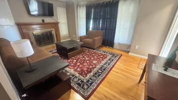 A living room with a fireplace, a television mounted on the wall, a brown armchair, a coffee table, and a patterned rug.