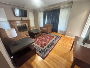 A living room with a fireplace, a television mounted on the wall, a brown armchair, a coffee table, and a patterned rug. - Welcome to Kings Landing (Detroit)