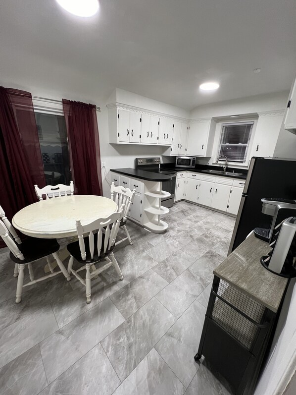 Een kleine keuken met witte kastjes, een magnetron, een spoelbak en een eethoek met een tafel en stoelen.