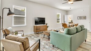 A modern living room with a sofa, a television, a dining area, and a ceiling fan. - 2BR Greer Condo: Pet-Friendly, King Beds, Fire Pit (Greer)