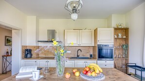 Una cocina con armarios blancos, encimera de madera, una mesa de comedor con frutas y un jarrón, y una cafetera.