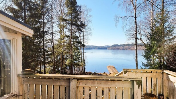 Una terraza de madera con vistas a un lago, rodeada de árboles densos y una orilla distante.