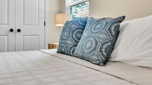 A neatly made bed with patterned pillows, a bedside lamp, and a window with blinds. - Greer Charm: Pet-Friendly 2BR with Fire Pit (Greer)