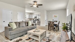 A modern living room with a gray sofa, a coffee table, and a dining area with a wooden table and chairs. A ceiling fan is visible, and the kitchen is in the background. - Greer Charm: Pet-Friendly 2BR with Fire Pit (Greer)