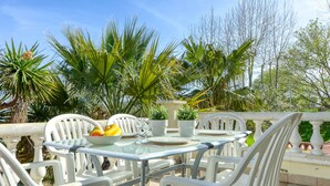 An outdoor dining area with a glass table and white chairs, surrounded by lush greenery and palm trees. - Apartment Muga by Interhome (Empuriabrava)