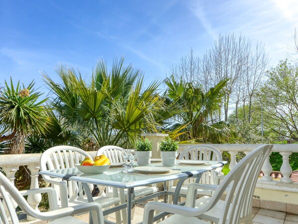 An outdoor dining area with a glass table and white chairs, surrounded by lush greenery and palm trees.