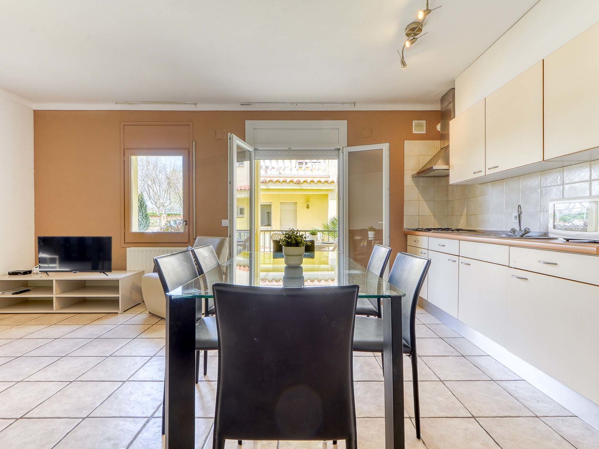 A modern kitchen with white cabinets, a dining table with four chairs, and a TV on a stand.