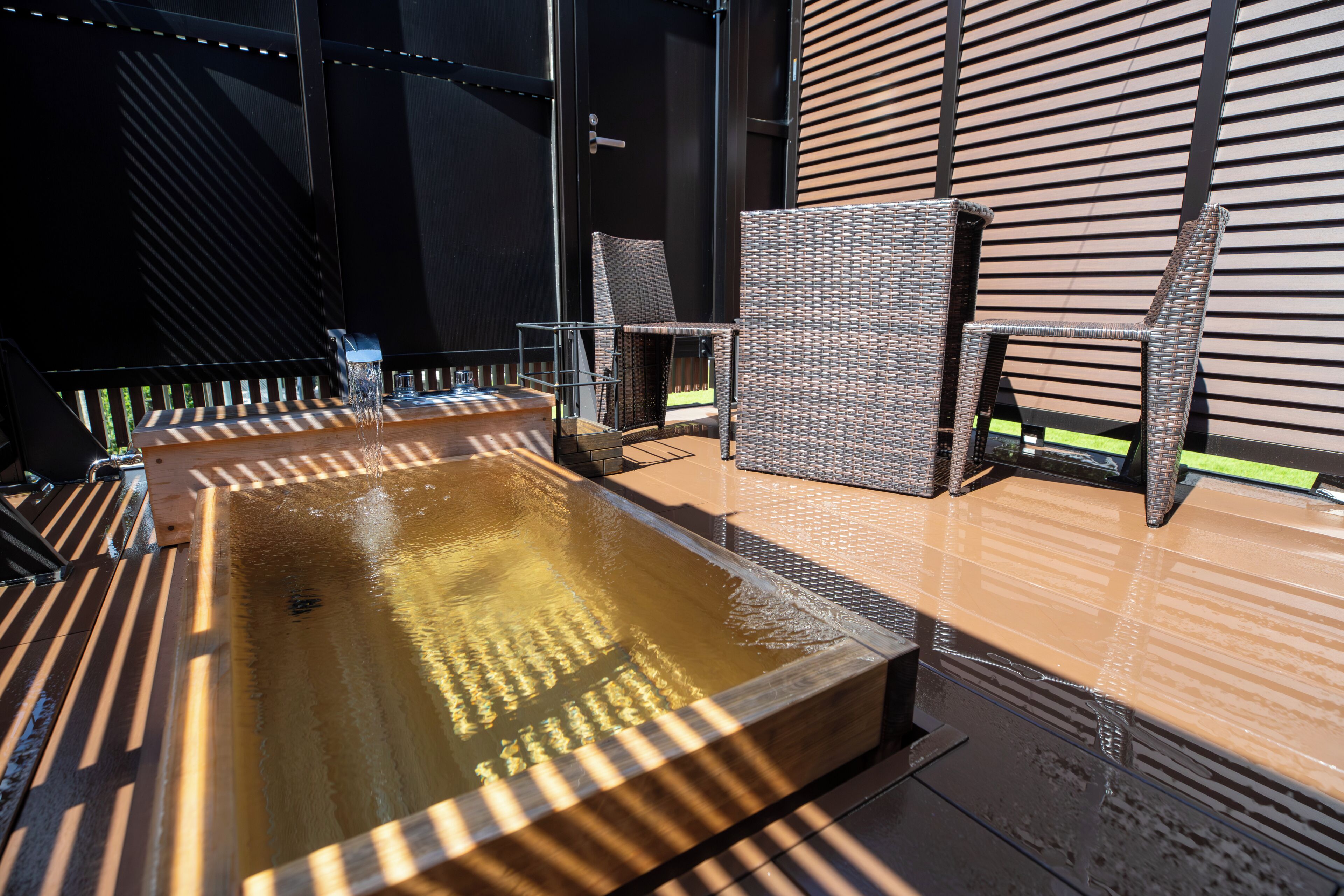 Un jacuzzi con sillones de mimbre, una terraza de madera y un edificio moderno con líneas horizontales al fondo.
