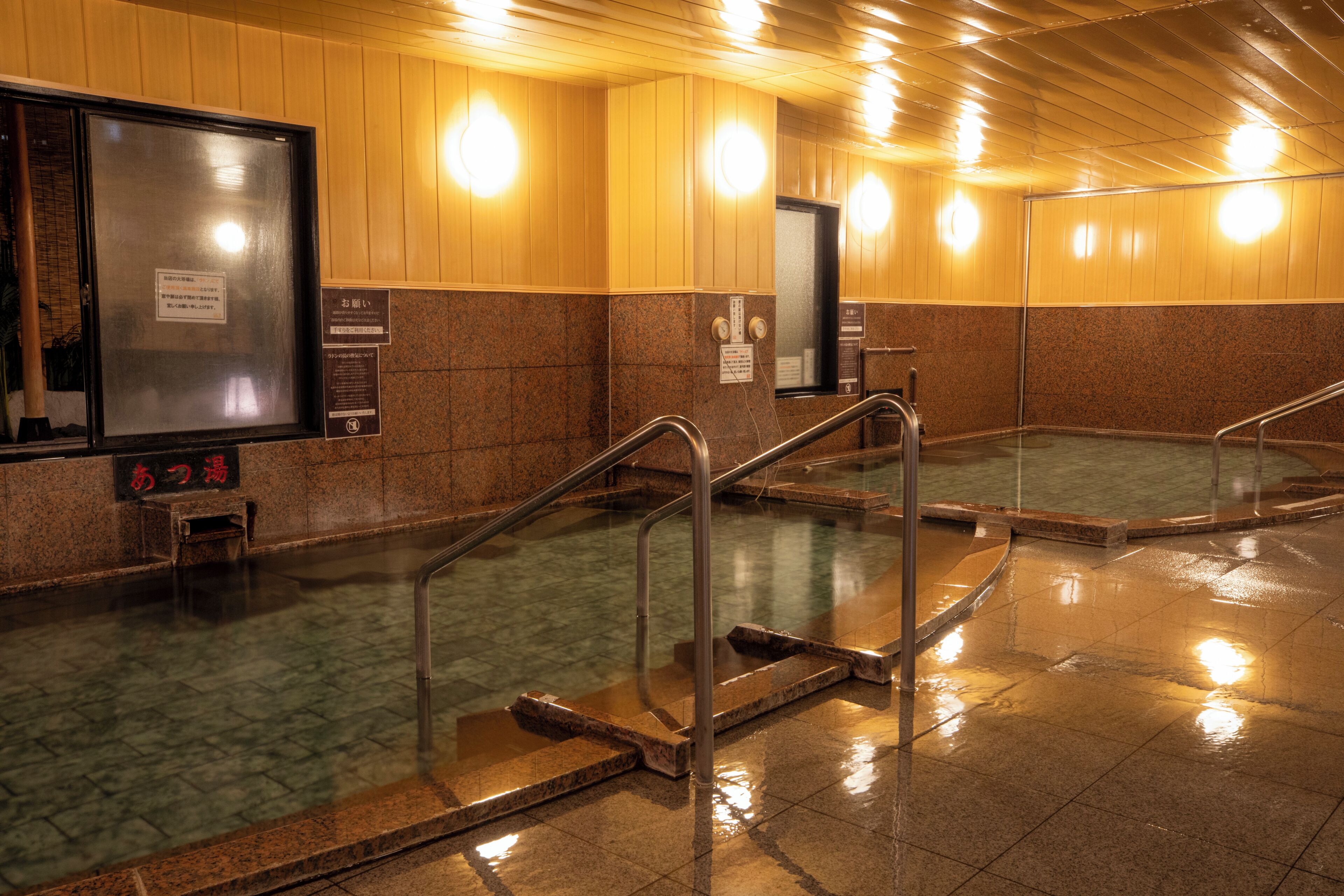 Un área de baños públicos con múltiples tinas de hidromasaje, barandas de metal y señalización informativa.