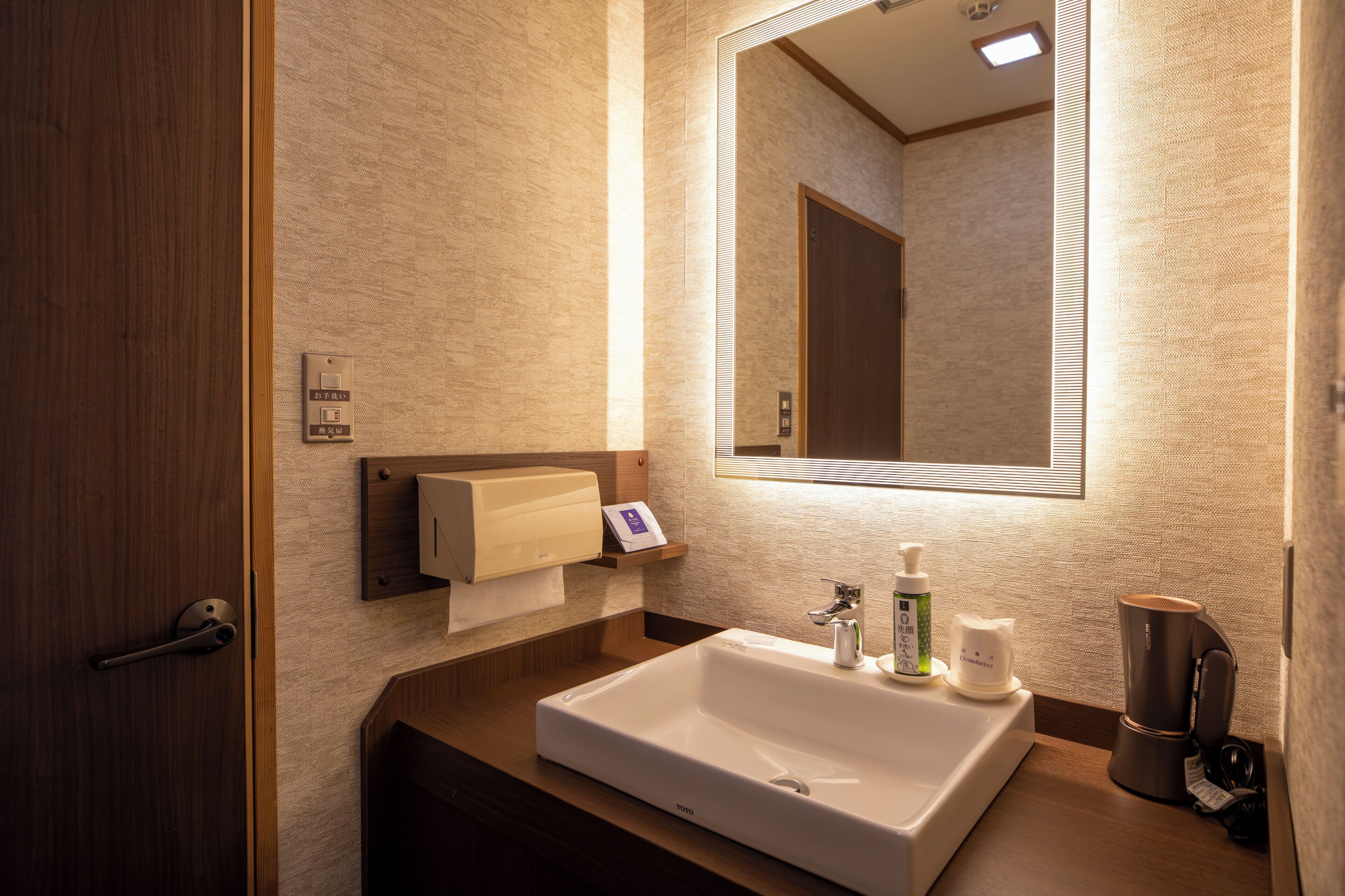 A hotel bathroom with a large mirror, a sink, a towel dispenser, and a soap dispenser.