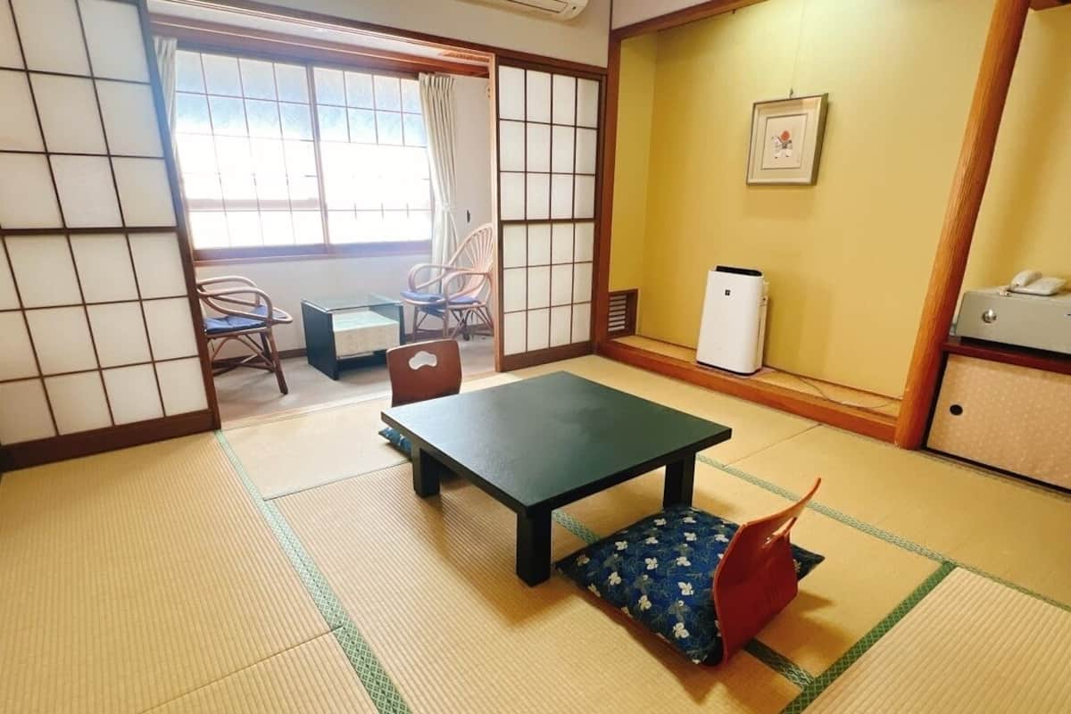 Ein Raum mit Tatami-Matten, einem niedrigen Tisch und einem Kissen. Eine Schiebetür führt zu einem Balkon mit Stühlen.