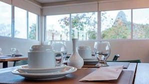 A dining table set with white dishes and glasses, with a view of greenery and a mountain through large windows. - Pousada Divino Morro (Fernando de Noronha)