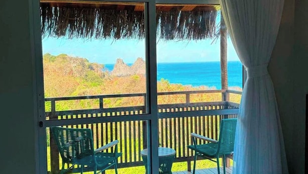 A balcony with two chairs and a small table, overlooking a beach and ocean. - Pousada Divino Morro (Fernando de Noronha)