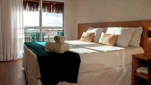 A neatly made bed with white linens and a green throw blanket, a wooden headboard, and a view of a balcony with green chairs. - Pousada Divino Morro (Fernando de Noronha)