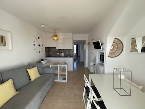 Een moderne woonkamer met een grijze bank, een eettafel met stoelen, een keuken op de achtergrond en een gang die naar een andere kamer leidt.