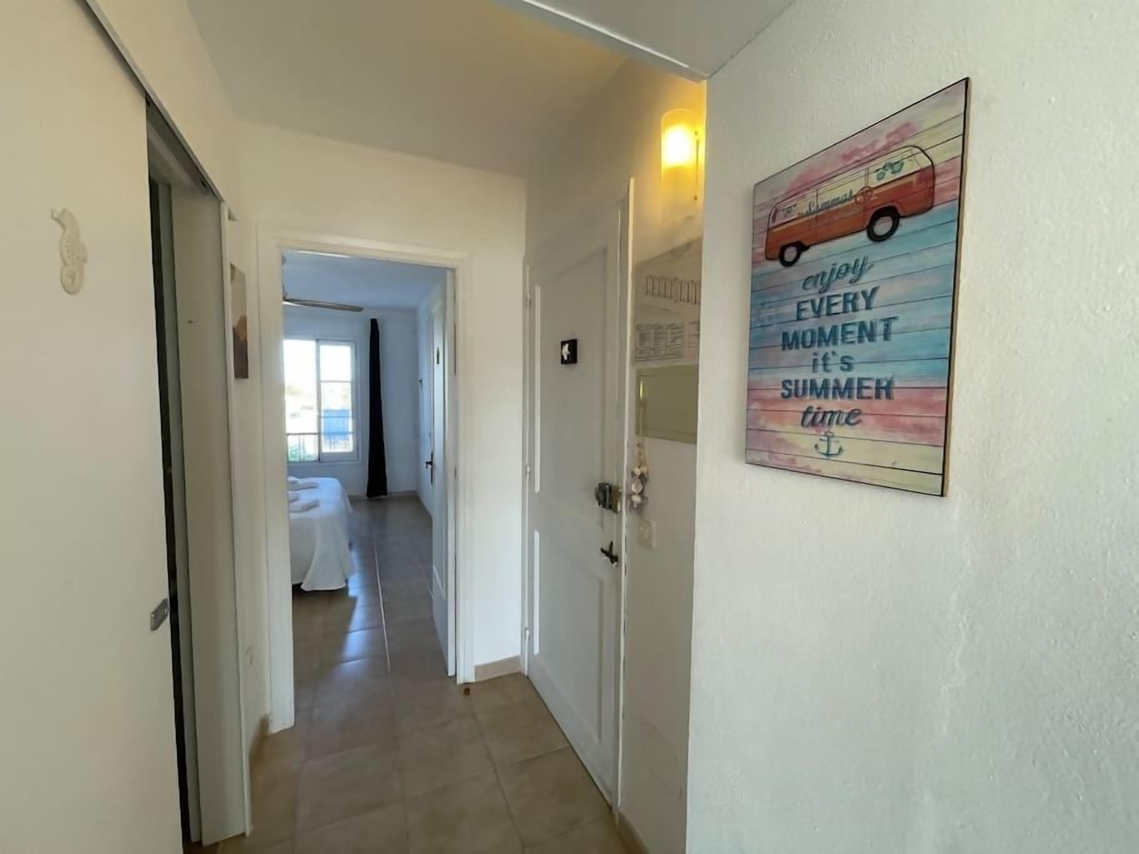 Een hal met een bed op de achtergrond, een deur links en een muur met een zomerposter.