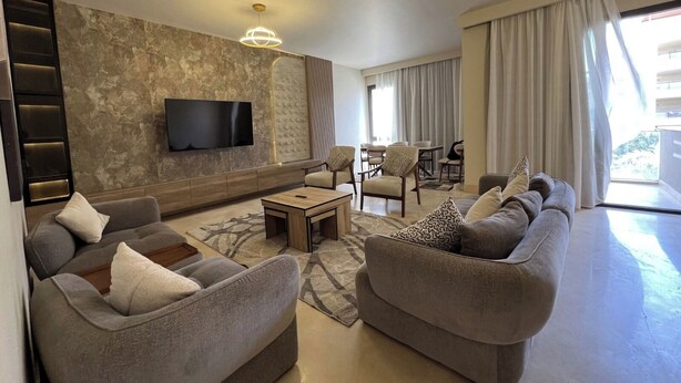 A modern living room with a flat-screen TV, grey sofas, a coffee table, and a dining area with chairs. - ALSAWAH 3 Bedroom Apartment at Allegria Sodic Sheikh Zayed (Sheikh Zayed City)