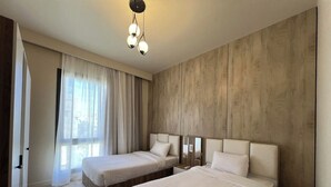 A hotel room with two beds, a wooden wall, a large window with curtains, and a hanging light fixture. - ALSAWAH 3 Bedroom Apartment at Allegria Sodic Sheikh Zayed (Sheikh Zayed City)